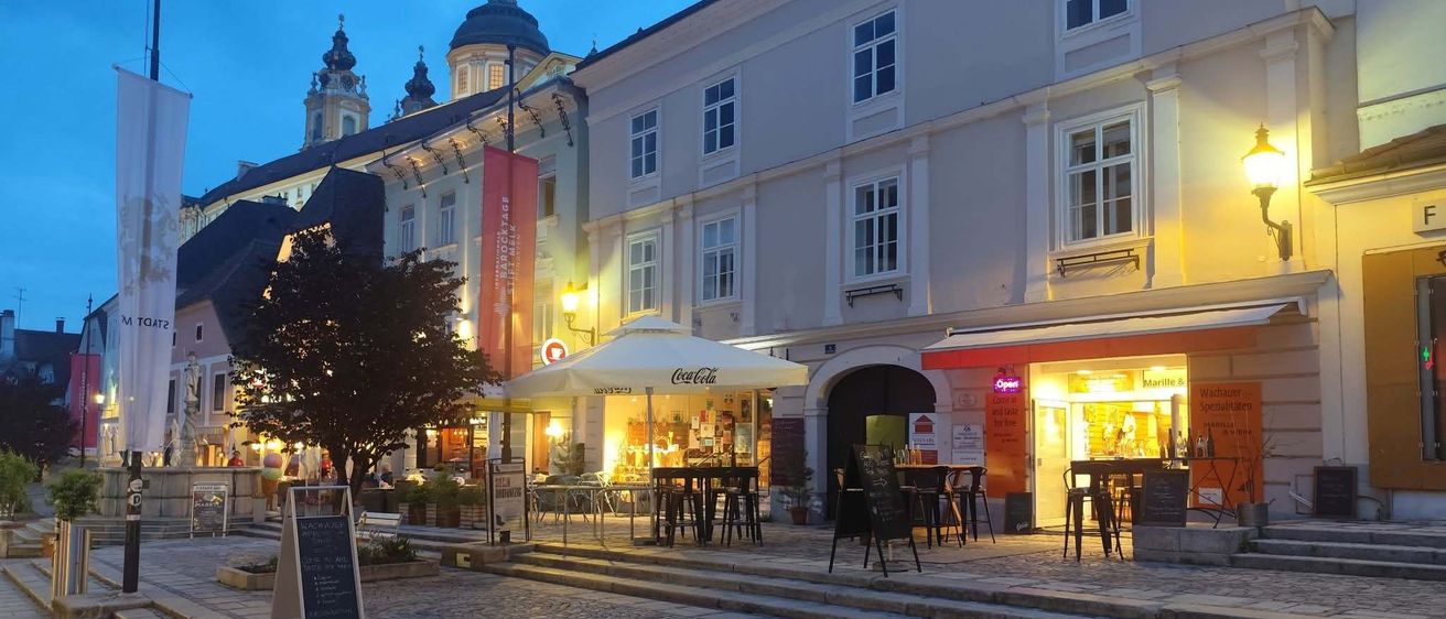Ein Outdoor-Café ist auf einer Kopfsteinpflasterstraße vor einem großen Gebäude mit mehreren Fenstern aufgestellt. Es ist Nacht, und es gibt mehrere Tische und Stühle unter einem Schirm.