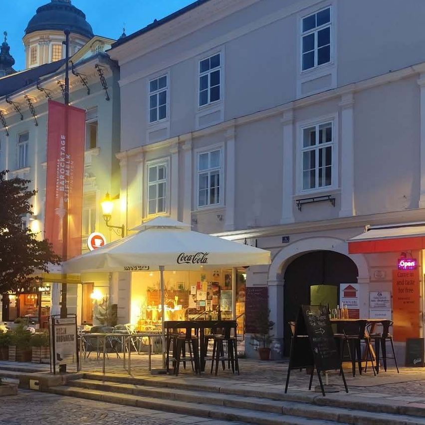 Ein Outdoor-Café ist auf einer Kopfsteinpflasterstraße vor einem großen Gebäude mit mehreren Fenstern aufgestellt. Es ist Nacht, und es gibt mehrere Tische und Stühle unter einem Schirm.