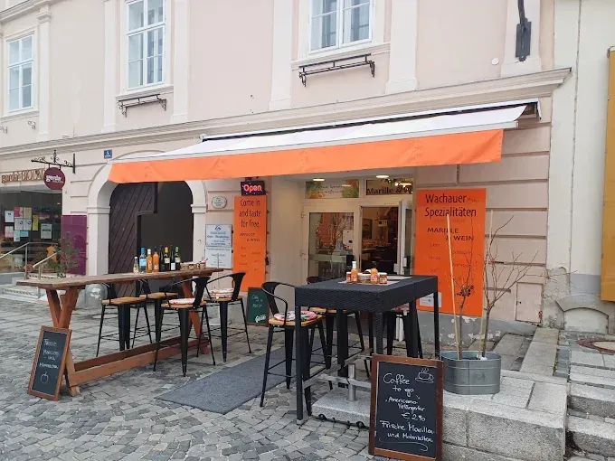 Outdoor seating at a coffee shop. Orange awning with white stripe. Tables with chairs and chalkboard menus. One table has wine bottles. The shop name is Marillie.