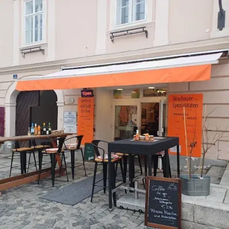 Terrasse eines Cafés. Oranger Vorbau mit weißem Streifen. Tische mit Stühlen und Kreidetafelmenüs. Ein Tisch hat Weinflaschen. Der Ladenname ist Marillie.