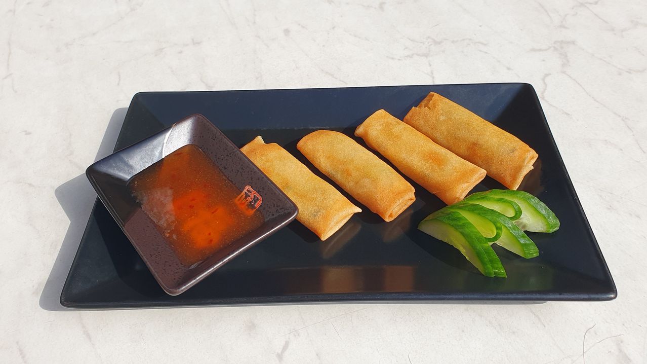 A black plate displays four fried spring rolls with a small square dish of dipping sauce and cucumber slices on the side.