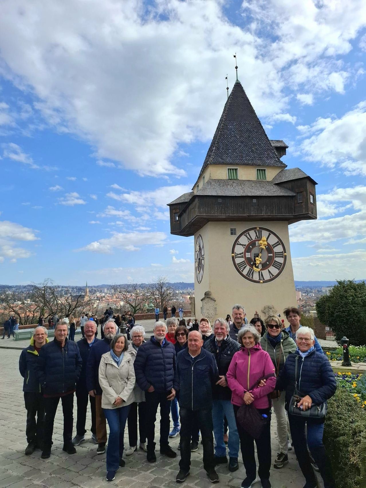 Eine Gruppe von Menschen, in kühleres Wetter gekleidet, posiert vor einem großen Uhrturm mit römischen Ziffern, vor dem Hintergrund eines klaren Himmels mit verstreuten Wolken und einer entfernten Stadtlandschaft.