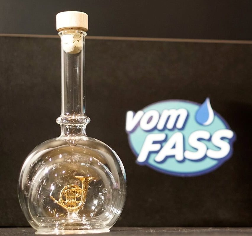 Eine klare Glasflasche mit einem goldenen französischen Horn im Inneren, einem Korken auf der Oberseite und einem vom FASS Logo im Hintergrund.