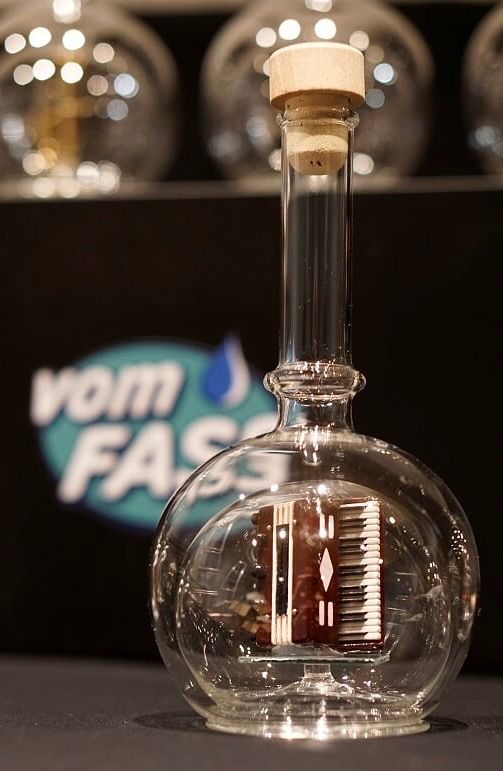 Eine durchsichtige Glasflasche mit einem Korkenverschluss enthält ein Miniatur-Akkordeon. Dahinter befindet sich ein schwarzer Hintergrund mit dem Wort 'VAM'. Im Hintergrund sind verschwommene Glasbehälter.