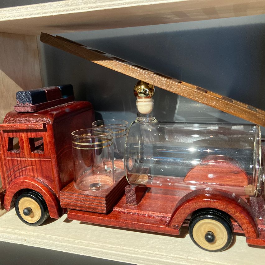Ein hölzerner Spielzeug-Löschwagen mit einer Glasflasche und zwei Gläsern auf einem hölzernen Regal. Die Flasche hat eine goldene Spitze.