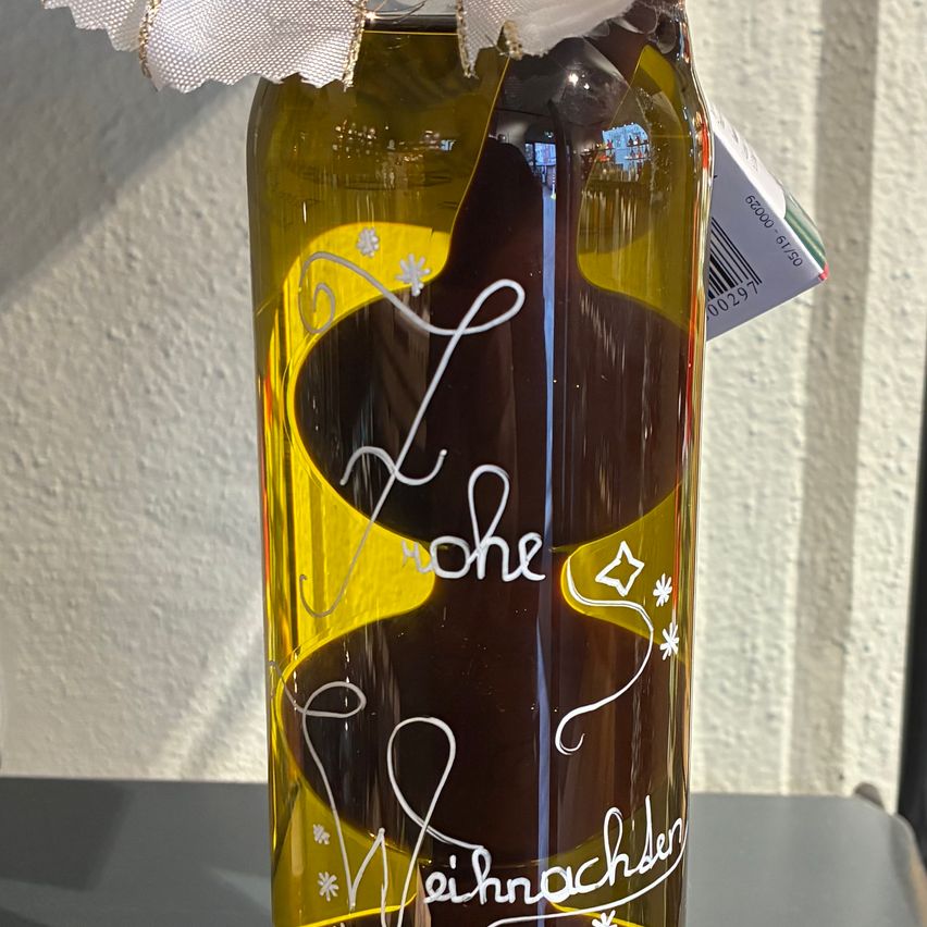 Eine Flasche mit dunkler Flüssigkeit, möglicherweise Olivenöl, mit gelbem Boden und weißen Verzierungen auf einem Tisch. Sie ist in ein weißes Band gewickelt.