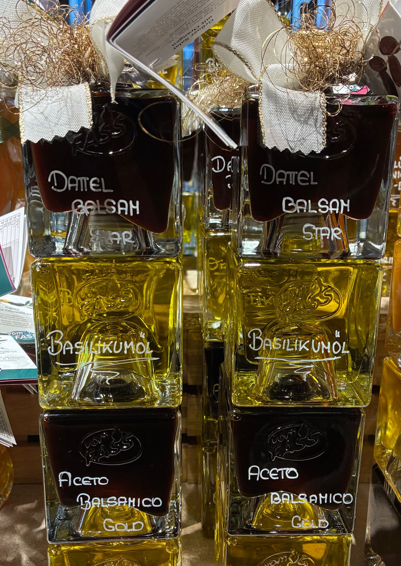 Zwei Flaschen Basilikumol und Dattel Balsam stehen auf einer Holzoberfläche. Beide sind in klaren Glasflaschen mit schwarzen Basen. Die Basilikumol-Flaschen haben gelbe Flüssigkeit und weißen Text, während die Dattel Balsam-Flaschen dunkle Flüssigkeit und weißen Text haben.