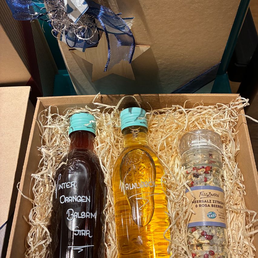 Eine Geschenkbox enthält drei Flaschen. Eine ist eine dunkle Flasche mit der Aufschrift Winter Orangen Balsam. Die zweite ist eine gelbe Flasche mit der Aufschrift Malvasia. Die dritte ist ein Glasgefäß mit der Aufschrift Meersalz Zitrone & Rosa Beeren.