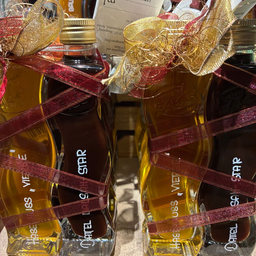 Drei Flaschen mit Flüssigkeit, jede mit dem Aufkleber 'USS VIETE', sind in rote Bänder gewickelt und mit goldenen Schleifen gebunden. Die Flüssigkeit in den Flaschen scheint dunkelbernsteinfarben zu sein.