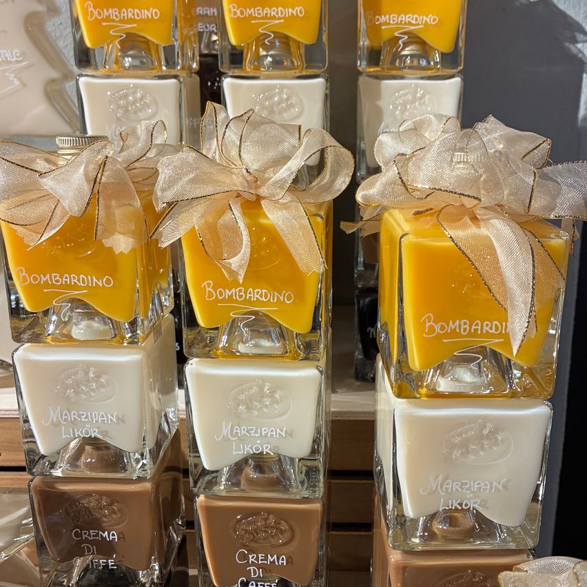 Eine Stapel von Glasbehältern mit gelbem, weißem und braunem Inhalt, jeweils mit 'Bombardino', 'Marzipan Likör' und 'Crema Di Caffè' beschriftet. Jeder Behälter ist mit einer Schleife geschmückt. Die Behälter sind ordentlich angeordnet.