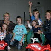 Eine Gruppe von Kindern und zwei Frauen posieren für ein Foto. Sie alle lächeln und haben ihre Hände erhoben. Die Kinder sitzen auf Spielzeugautos und eine Frau hält ein Baby. Die Umgebung scheint drinnen zu sein.