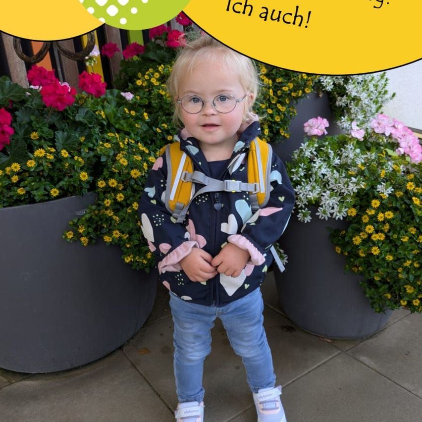 Ein kleines Mädchen mit Brille und einem Rucksack steht vor bunten Blumen und einer gelben Sprechblase, die 'Ich auch!' sagt.