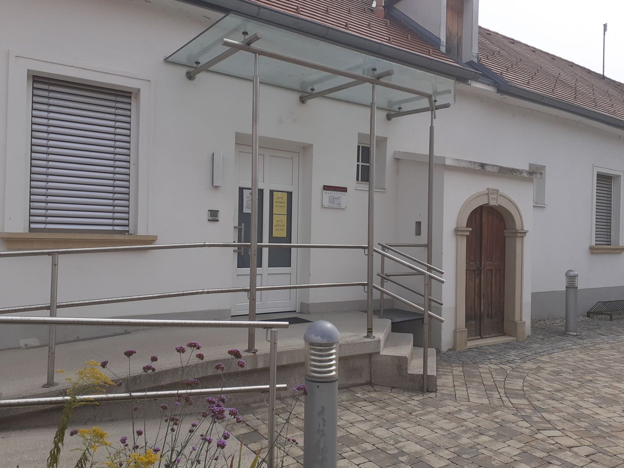 Ein weißes Gebäude mit einem Glasvordach über dem Eingang, Edelstahl-Handläufen und einer Holztür mit einem Schild darüber.