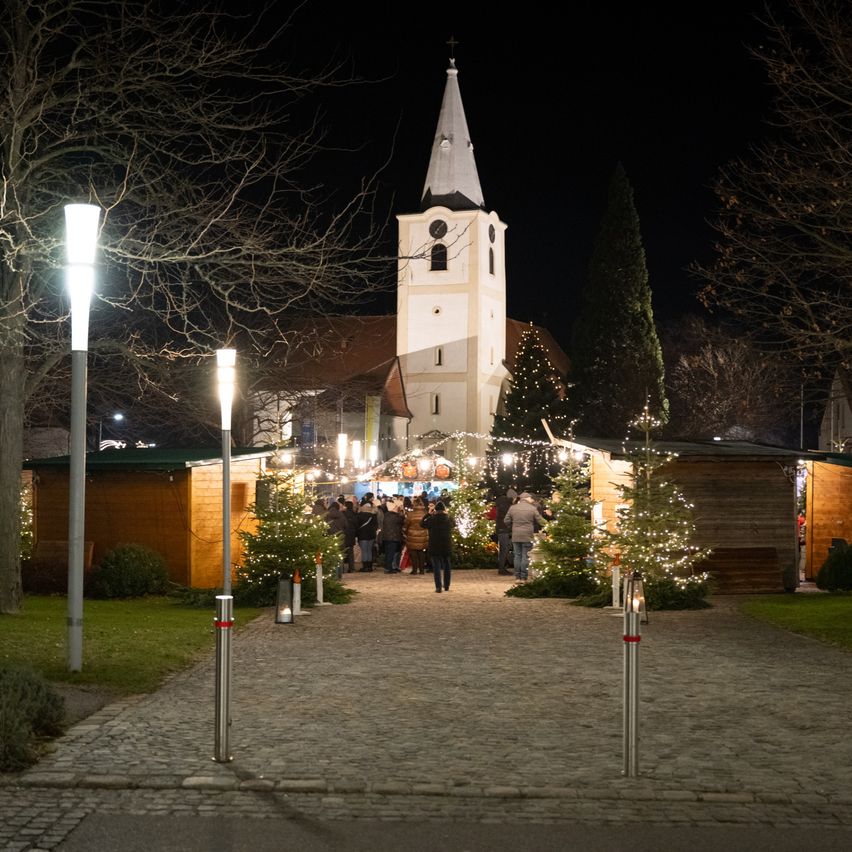 Ein nächtlicher Blick auf eine Kirche mit einem Kirchturm, umgeben von Weihnachtsbäumen und Menschen. Es gibt Stände und brennende Kerzen.