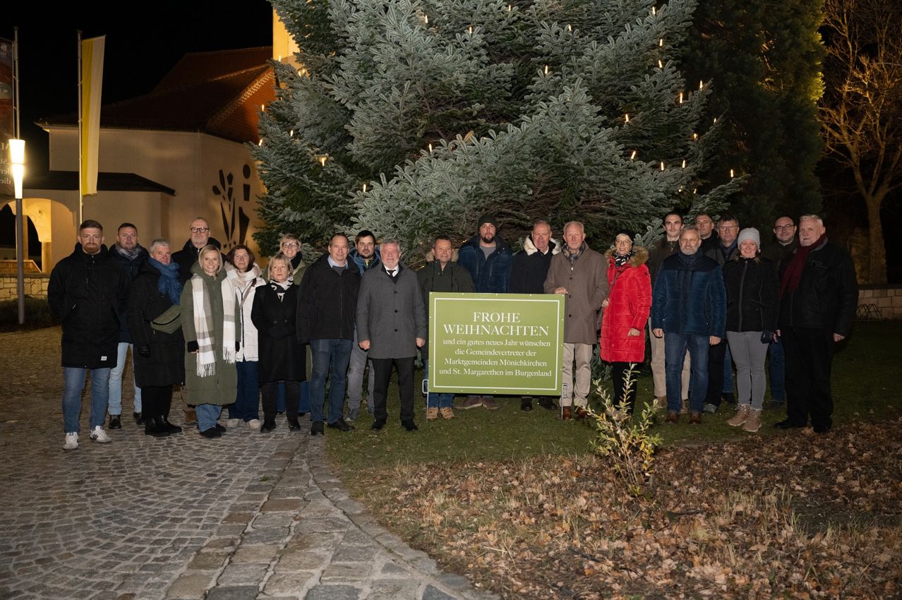 Eine Gruppe von Menschen steht vor einem großen geschmückten Weihnachtsbaum mit einem Schild, das 'Frohe Weihnachten' steht. Das Foto wurde bei Nacht aufgenommen.