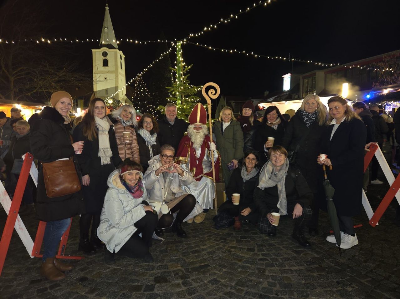 Eine Gruppe von Menschen posiert für ein Foto mit dem Weihnachtsmann vor einem geschmückten Weihnachtsbaum, der von Lichterketten erhellt wird, in der Nähe eines Kirchturms bei Nacht.