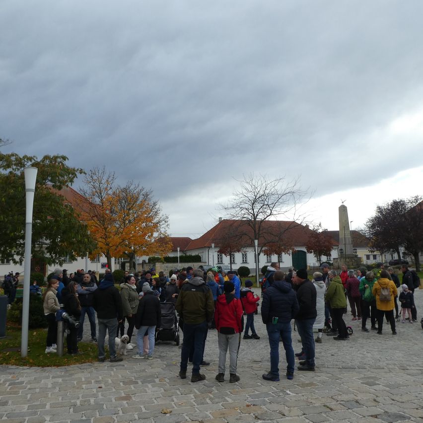 Eine Menschenmenge versammelt sich auf einem gepflasterten Platz, einige tragen Mützen und Jacken, mit Gebäuden, Bäumen und einem Denkmal im Blick. Der Himmel ist bewölkt.