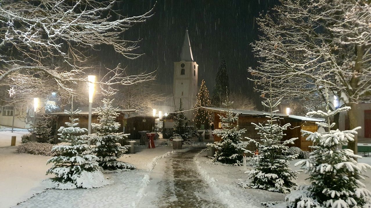 Eine verschneite Nachtszene zeigt eine Kirche mit einem Kirchturm, geschmückt mit einem Weihnachtsbaum. Ein Weg führt zur Kirche, gesäumt von schneebedeckten Bäumen und einer Straßenlaterne.