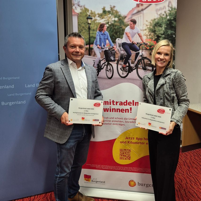 Ein Mann und eine Frau stehen lächelnd da und halten Urkunden vor einem Plakat von Radfahrern und einer großen Werbung. Das Plakat zeigt Text und Bilder von Menschen, die auf einer Straße Fahrrad fahren.