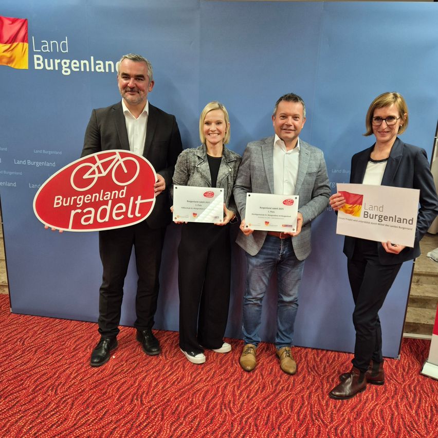 Vier Menschen stehen auf einem roten Teppich und lächeln. Sie halten Urkunden und ein Schild mit der Aufschrift 'Burgenland radelt'. Hinter ihnen ist eine Kulisse mit den Worten 'Land Burgenland' und einer Flagge.