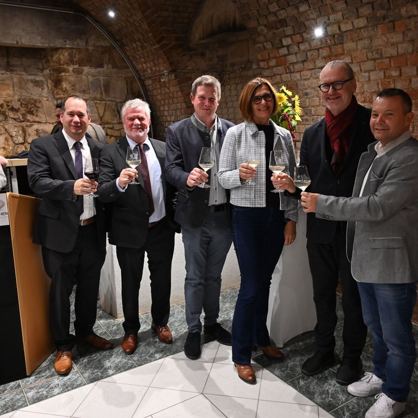 Eine Gruppe von Menschen steht in einem Raum mit Ziegelwänden, lächelt und hält Weingläser. Sie sind in formeller Kleidung gekleidet.
