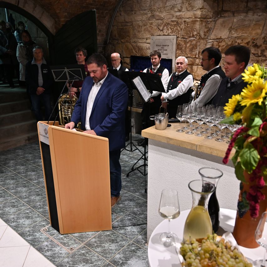 Ein Mann in einem blauen Anzug steht an einem Podium und spricht zu einer Gruppe von Musikern und Gästen. Die Musiker halten Blech- und Holzblasinstrumente. Auf dem Tisch stehen Weingläser, Blumen und Trauben.