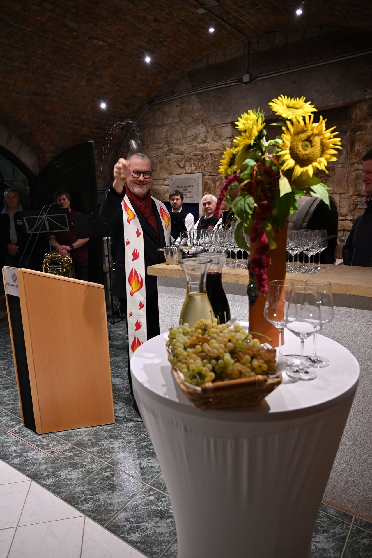 Ein Priester steht an einem Tisch mit einem Korb Trauben und Weingläsern. Hinter ihm spielt ein Musiker eine Tuba. Ein Strauß Sonnenblumen und eine Karaffe Wein stehen auf dem Tisch.