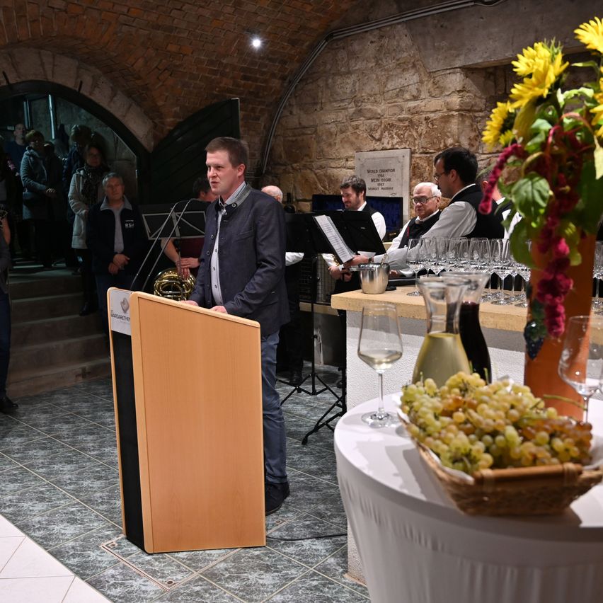 Ein Mann spricht an einem Podium in einem Raum mit Steinwänden, einer Messingtuba und Weingläsern. Andere Leute sind im Raum, einige sitzen und einige stehen.