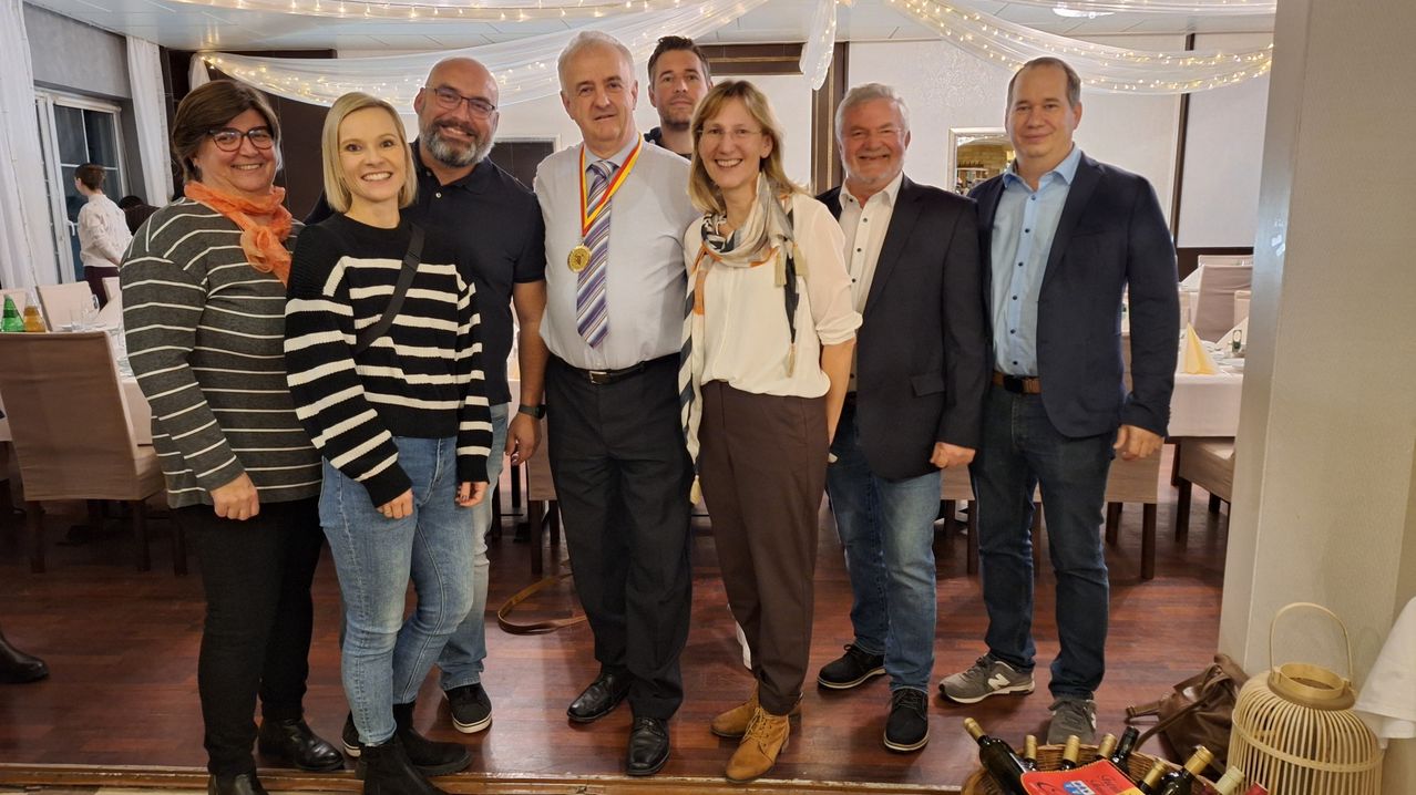 Eine Gruppe von sieben Personen posiert für ein Foto in einem Raum mit Holzboden. Der Mann in der Mitte trägt eine Medaille. Die Frau neben ihm lächelt. Der andere Mann trägt eine Uhr. Hinter ihnen befindet sich ein Spiegel mit der Reflexion eines Raumes.