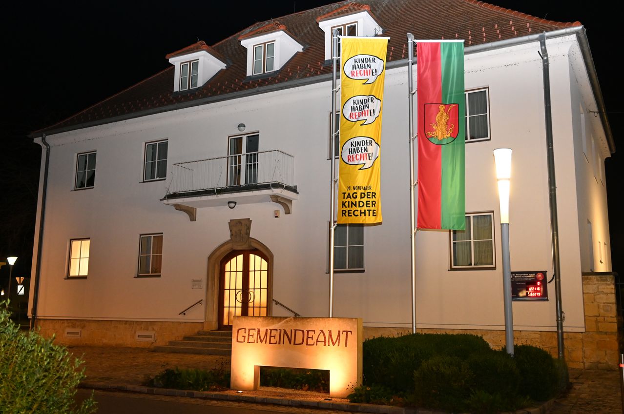 Ein Gebäude mit einem Banner, das 'Kinder haben Rechte' besagt, in der Nacht beleuchtet. Zwei Fahnen stehen neben dem Gebäude an Stangen, eine mit einem Logo und die andere mit '20. November' darauf. Das Gebäude hat ein Schild mit der Aufschrift 'Gemeindeamt'.