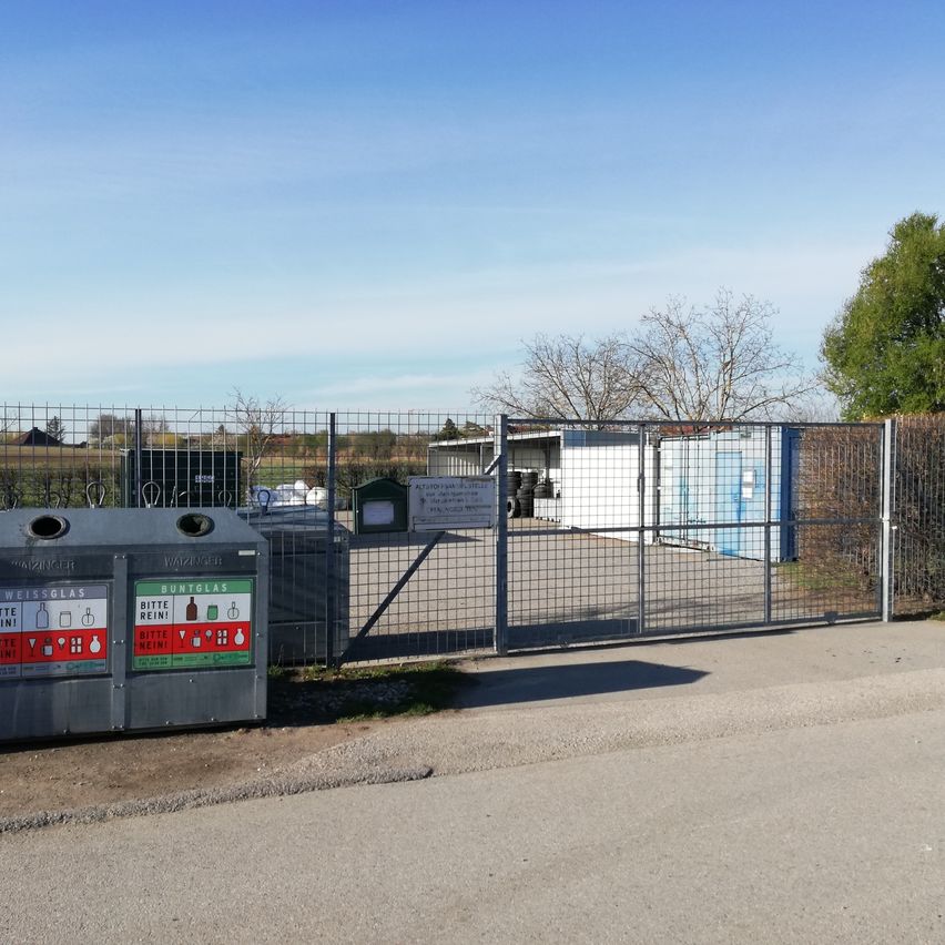 Ein Recyclingzentrum mit verschiedenen Containern, einschließlich eines für Glas, hinter einem Metallzaun. Das Zentrum ist von einem Betonweg und einigen Bäumen umgeben.