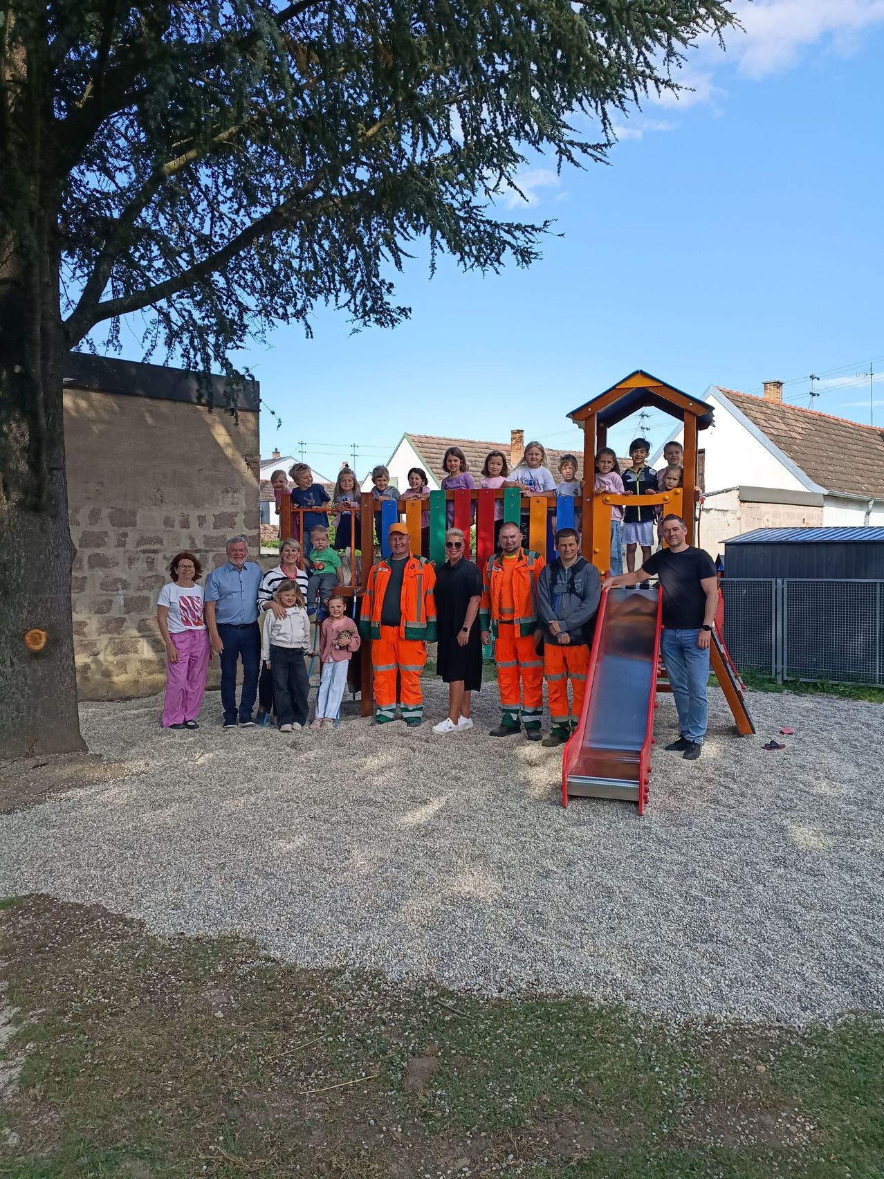 Eine Gruppe von Erwachsenen und Kindern posiert für ein Foto vor einem Spielplatz, wobei einige Arbeiter in Sicherheitsuniformen in der Nähe stehen.
