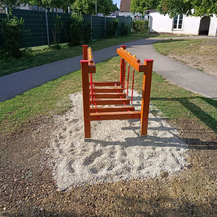 Eine hölzerne Kletterstruktur steht in einem Spielplatz. Sie ist von Kies und Gras umgeben. Ein Weg und ein grüner Zaun sind in der Nähe.