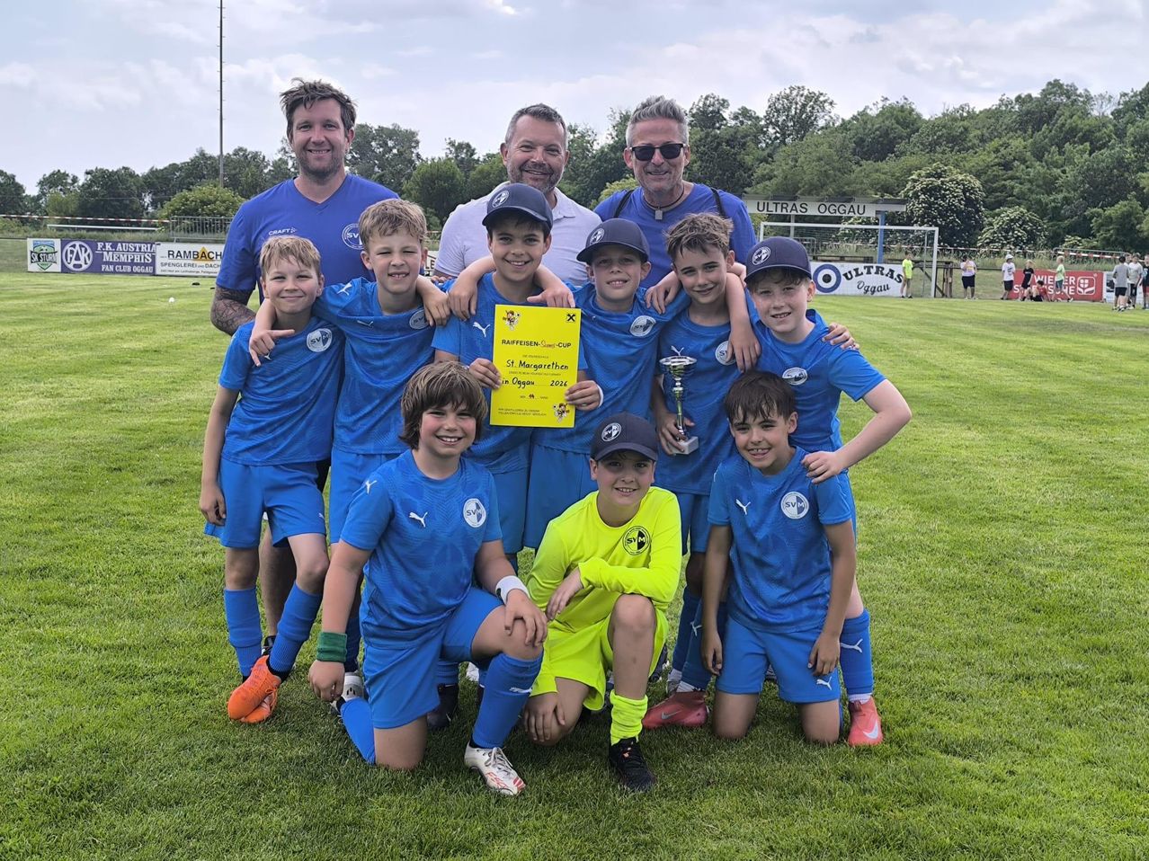 Eine Fußballmannschaft junger Jungen und ihrer Trainer posiert für ein Foto mit einem Pokal und einem Zertifikat an einem sonnigen Tag.