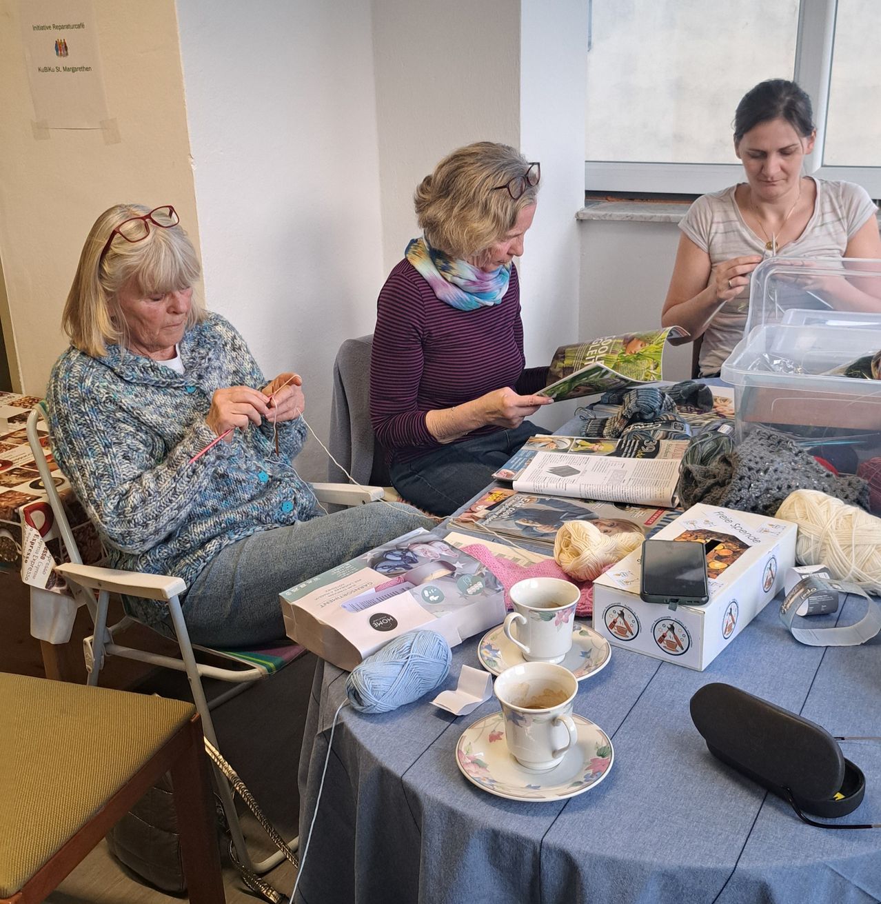Drei Frauen in einem Raum stricken. Die Frau links sitzt auf einem Stuhl. Die Frau in der Mitte sitzt auf einem Stuhl mit einem Tisch vor ihr. Die Frau rechts sitzt auf einem Stuhl und strickt.