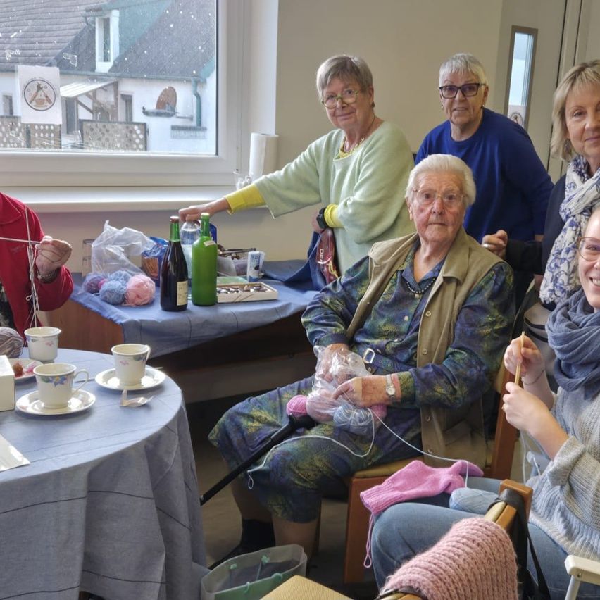 Eine Gruppe älterer Frauen strickt und trinkt Tee in einem Raum mit Fenster. Flaschen, Tassen und Garn liegen auf dem Tisch. Eine Frau benutzt einen Gehstock.