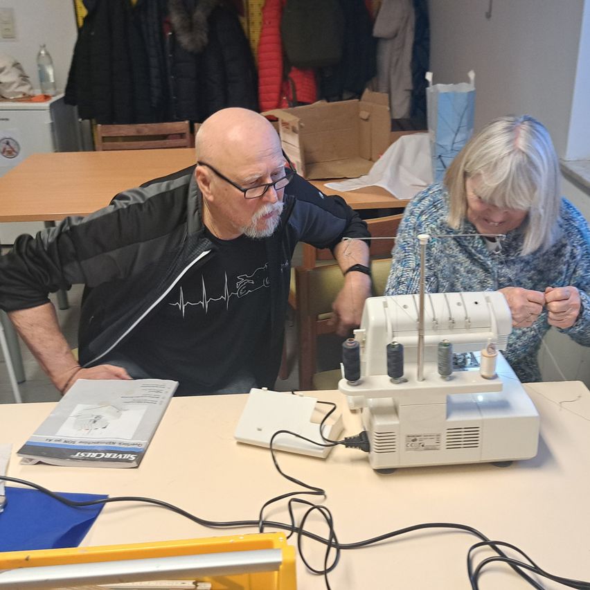 Ein älterer Mann und eine ältere Frau sitzen neben einer Nähmaschine, der Mann lehnt sich über sie, während die Frau lächelt und mit der Maschine arbeitet. Hinter ihnen sind Stühle und ein Tisch mit Jacken und einer Kiste zu sehen.