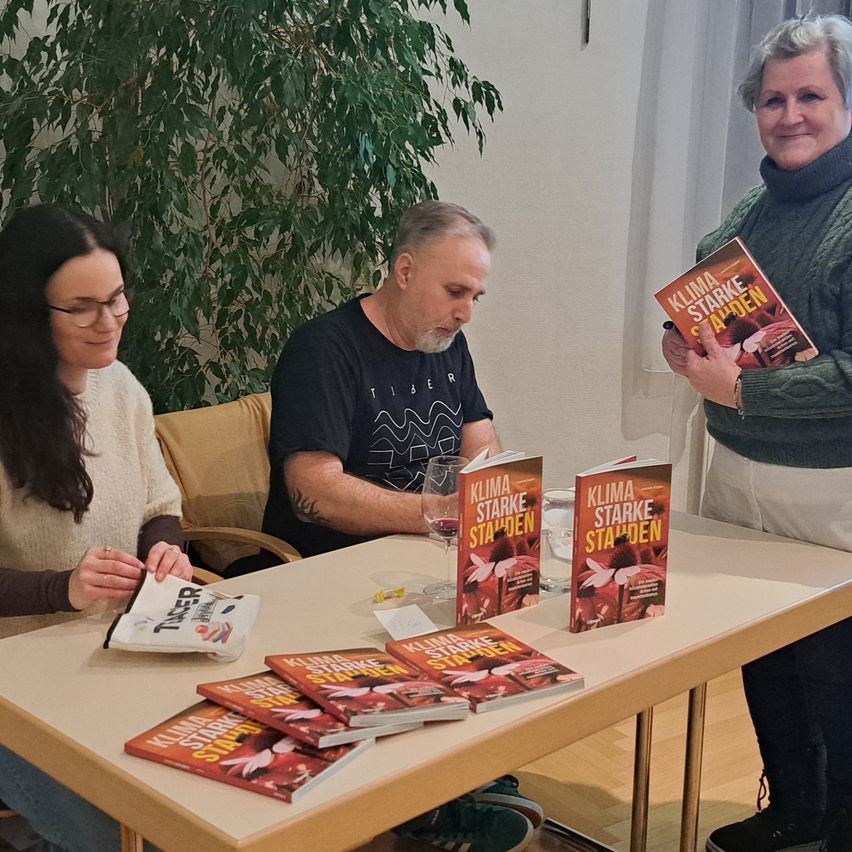Drei Personen versammeln sich um einen Tisch, einer hält ein Glas Wein, eine andere hält ein Buch mit dem Titel 'Klima Starke' und die dritte hält einen Stift.