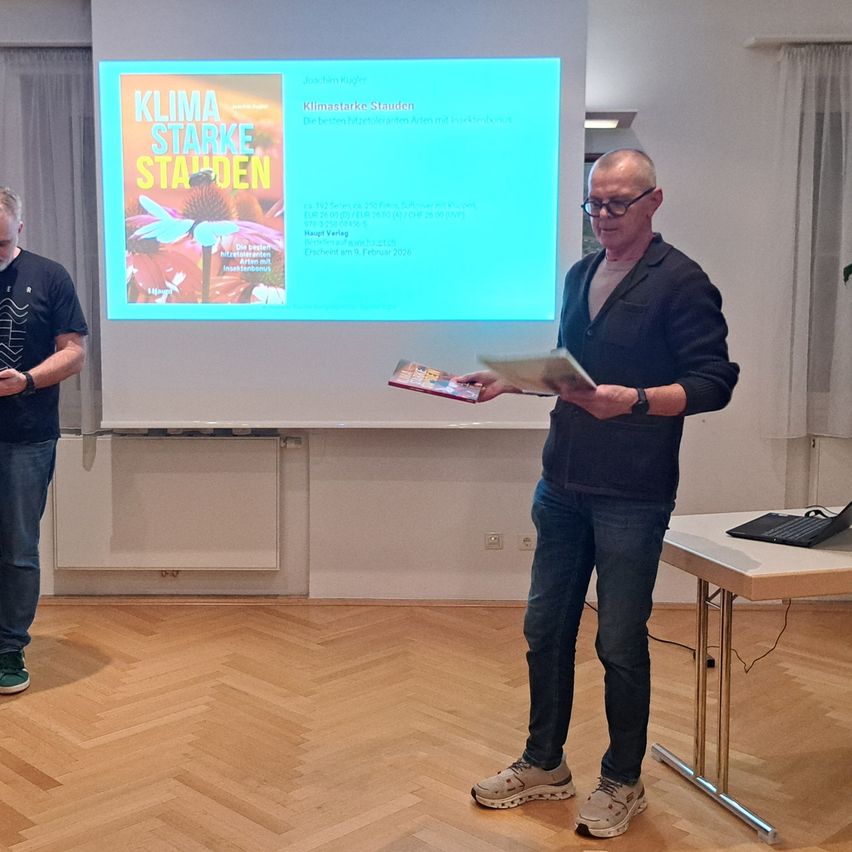 Ein Mann steht in einem Raum, hält ein Buch und liest, während ein anderer Mann links von ihm steht. Eine Projektionsleinwand zeigt ein Buchcover mit dem Titel 'Klima Starke Stadten'. Ein Schreibtisch mit Laptop steht rechts.