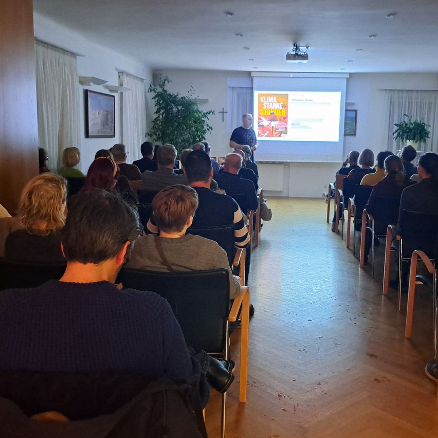 Ein Raum mit vielen Menschen, die auf Stühlen sitzen und einem Mann zuhören, der vor einer Projektionsleinwand steht.