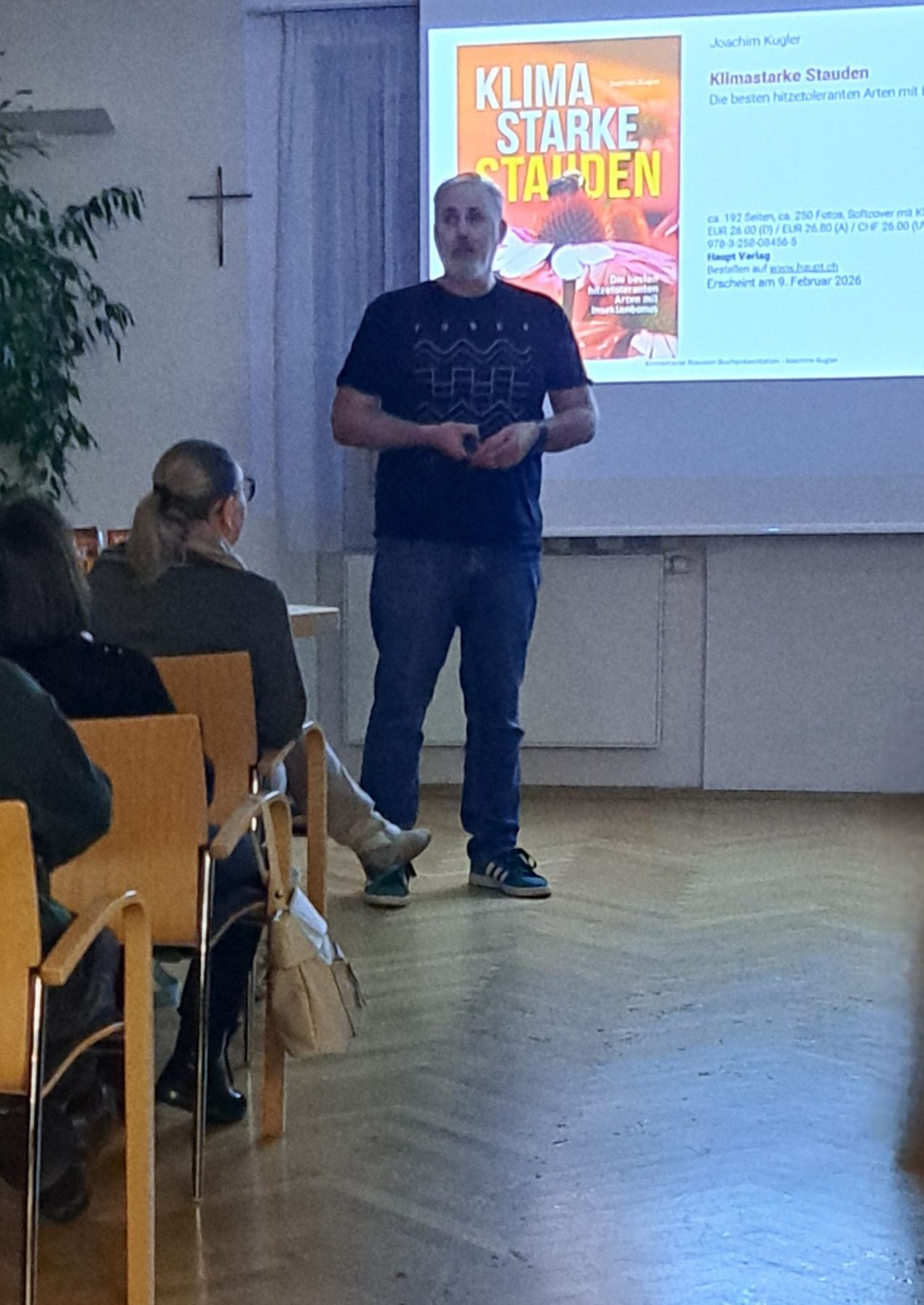 Ein Mann steht vor einer Projektionsleinwand und spricht zu einer Gruppe von Menschen, die auf Stühlen sitzen. Die Leinwand zeigt Informationen über eine Veranstaltung.