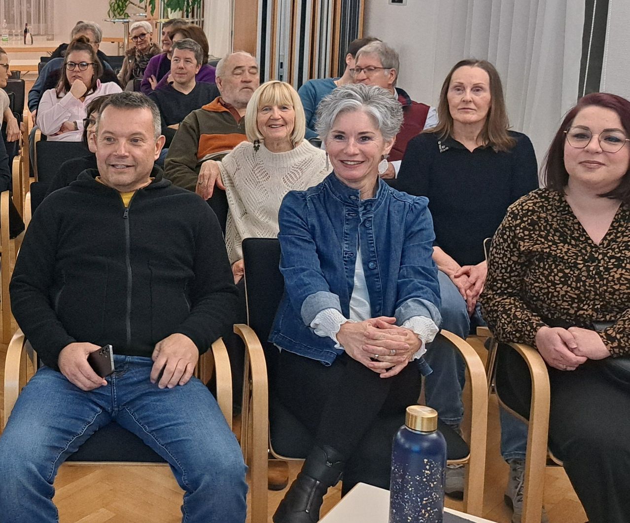 Eine Gruppe von Erwachsenen, sowohl Männer als auch Frauen, sitzen in Stühlen, manche lächeln, andere konzentrieren sich auf etwas. Ein Mann in einer schwarzen Jacke hält ein Telefon, während eine Frau in einer blauen Jacke einen Ring trägt. Eine blaue Flasche steht auf einem Tisch vor ihnen.