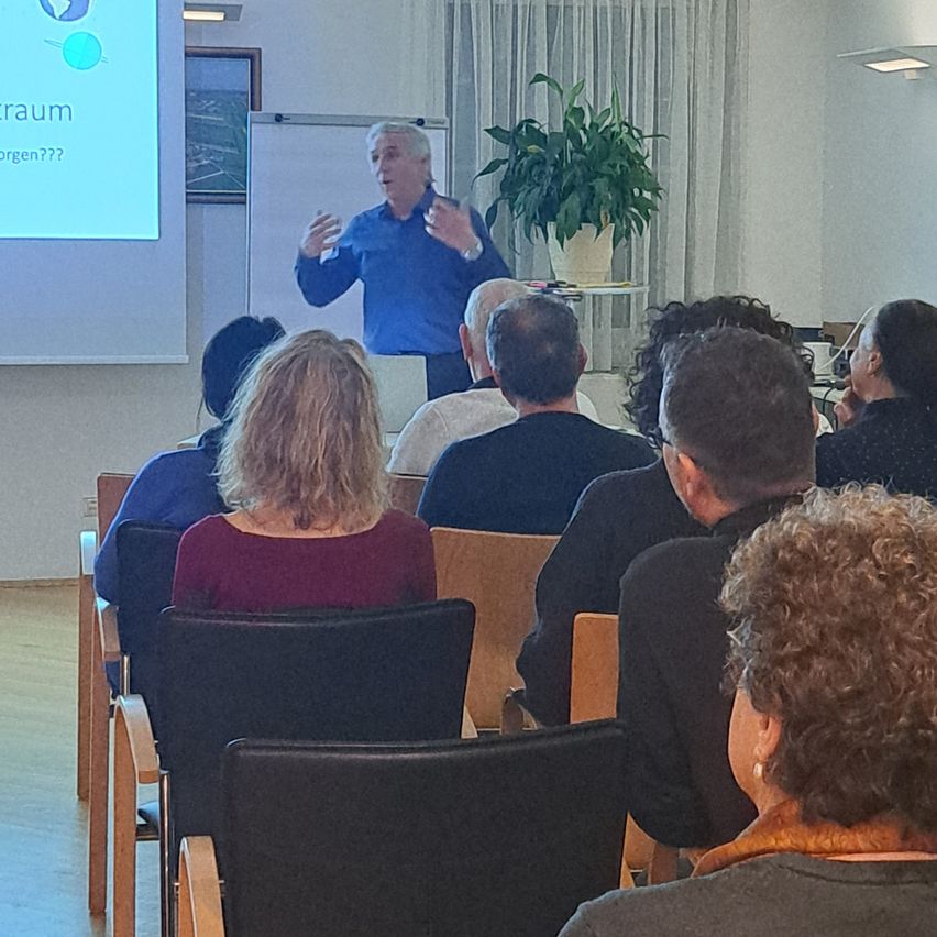 Ein Mann hält in einem Konferenzraum eine Präsentation vor einer Gruppe von Menschen. Das Publikum sitzt in Stühlen und hört aufmerksam zu.