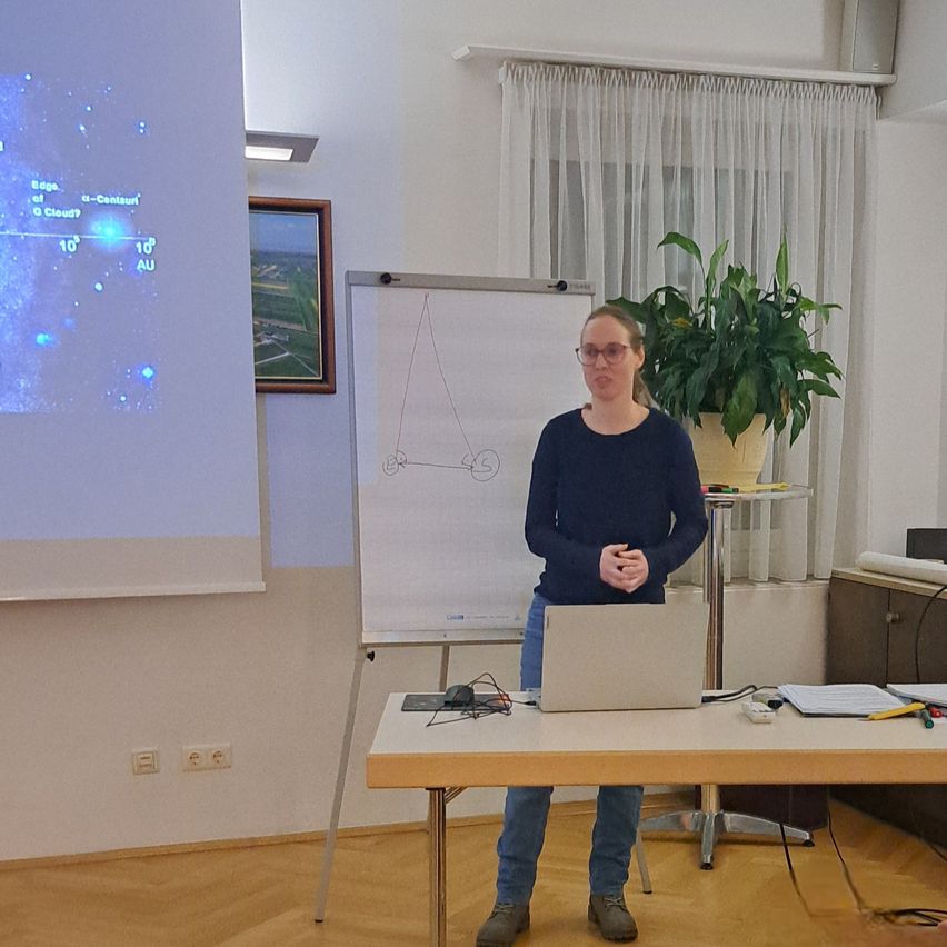 Eine Frau steht vor einem Tisch mit einem Laptop und hält eine Präsentation. Hinter ihr ist eine Tafel sichtbar, und eine Projektionsleinwand zeigt ein Bild des Himmels. Ein Topf mit einer Pflanze steht auf einem Tisch zu ihrer Rechten.