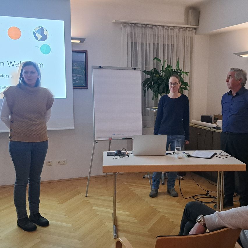Eine Frau steht vor einer Projektionsleinwand, während zwei andere hinter einem Tisch mit Laptop, Papieren und einem Glas Wasser stehen. Ein Mann steht in der Nähe und eine weitere Person sitzt auf einem Stuhl.