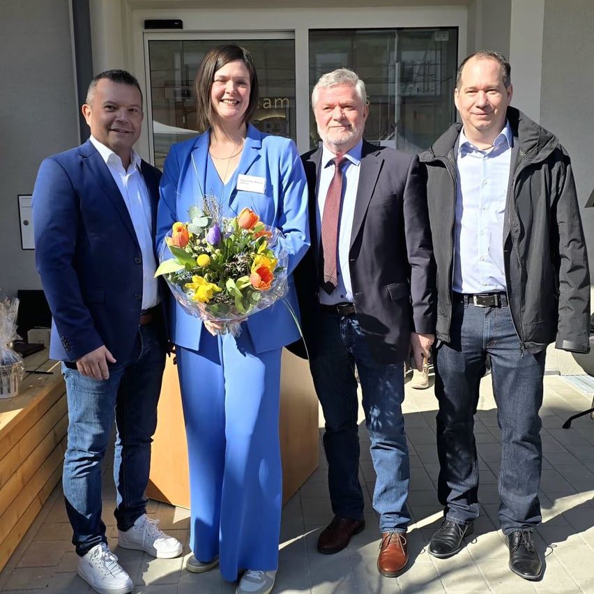 Vier Menschen stehen vor einem Gebäude, lächeln und posieren für ein Foto. Eine Frau in der Mitte hält einen Blumenstrauß. Hinter ihnen befindet sich eine Glastür mit einem Schild.