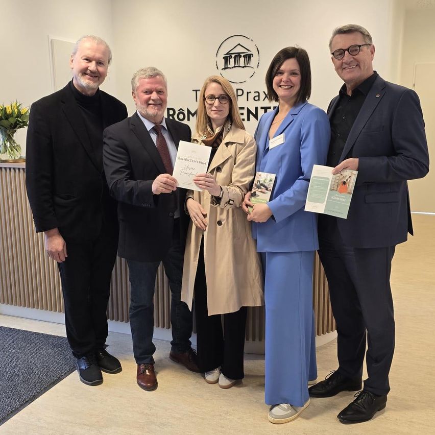 Fünf Personen in formeller Kleidung stehen nebeneinander, halten Urkunden und Broschüren und lächeln vor einer Wand mit dem Text 'Praxis'.