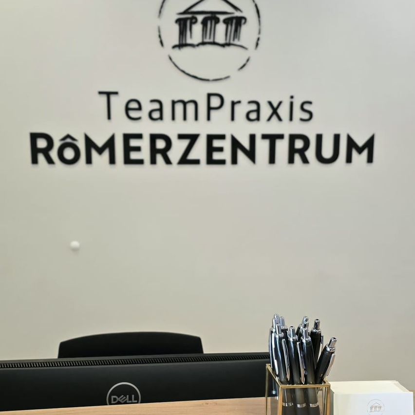 Ein Büro mit einem TeamPraxis Romerzentrum-Logo an einer weißen Wand, einem Schreibtisch und einem Dell-Monitor mit Stiften in einem Halter.