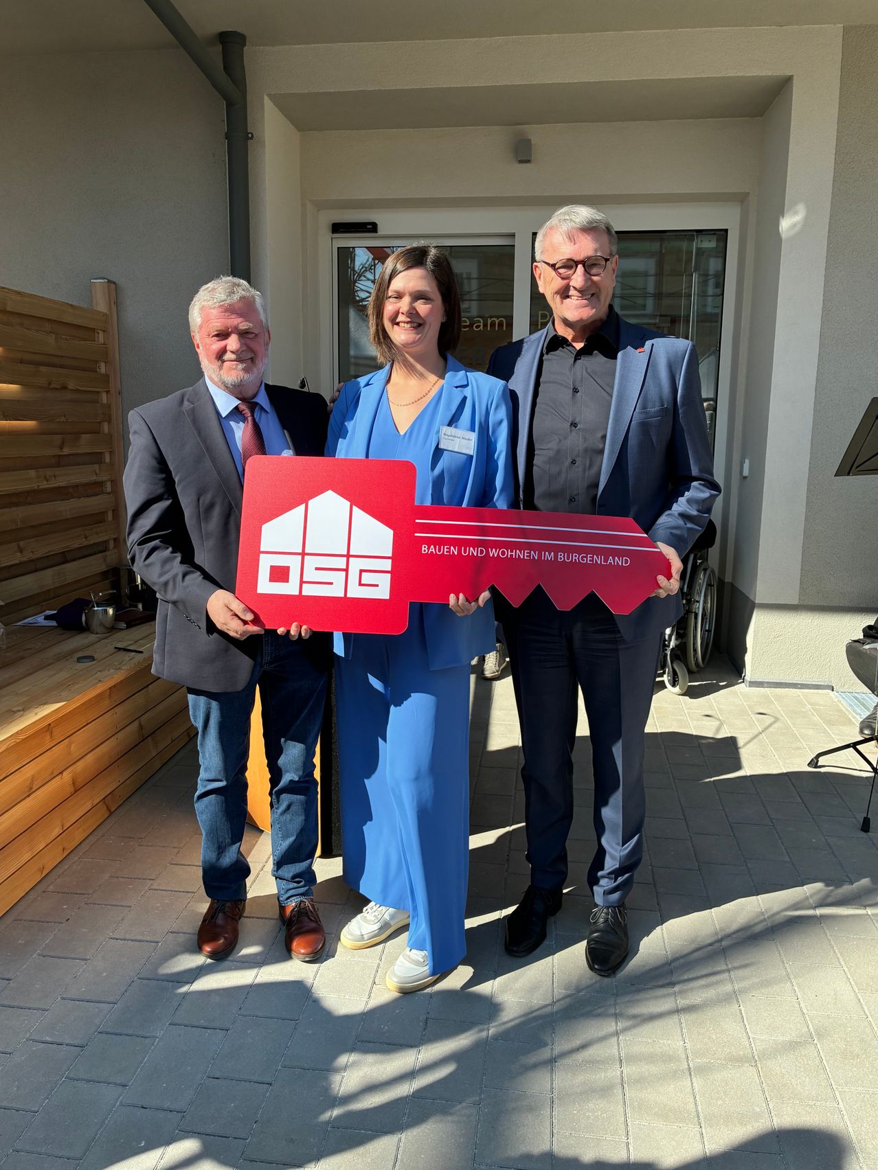 Drei Menschen stehen lächelnd vor einem Gebäude. Sie halten ein rotes Schlüsselsymbol mit der Aufschrift Bauen und Wohnen im Burgenland.