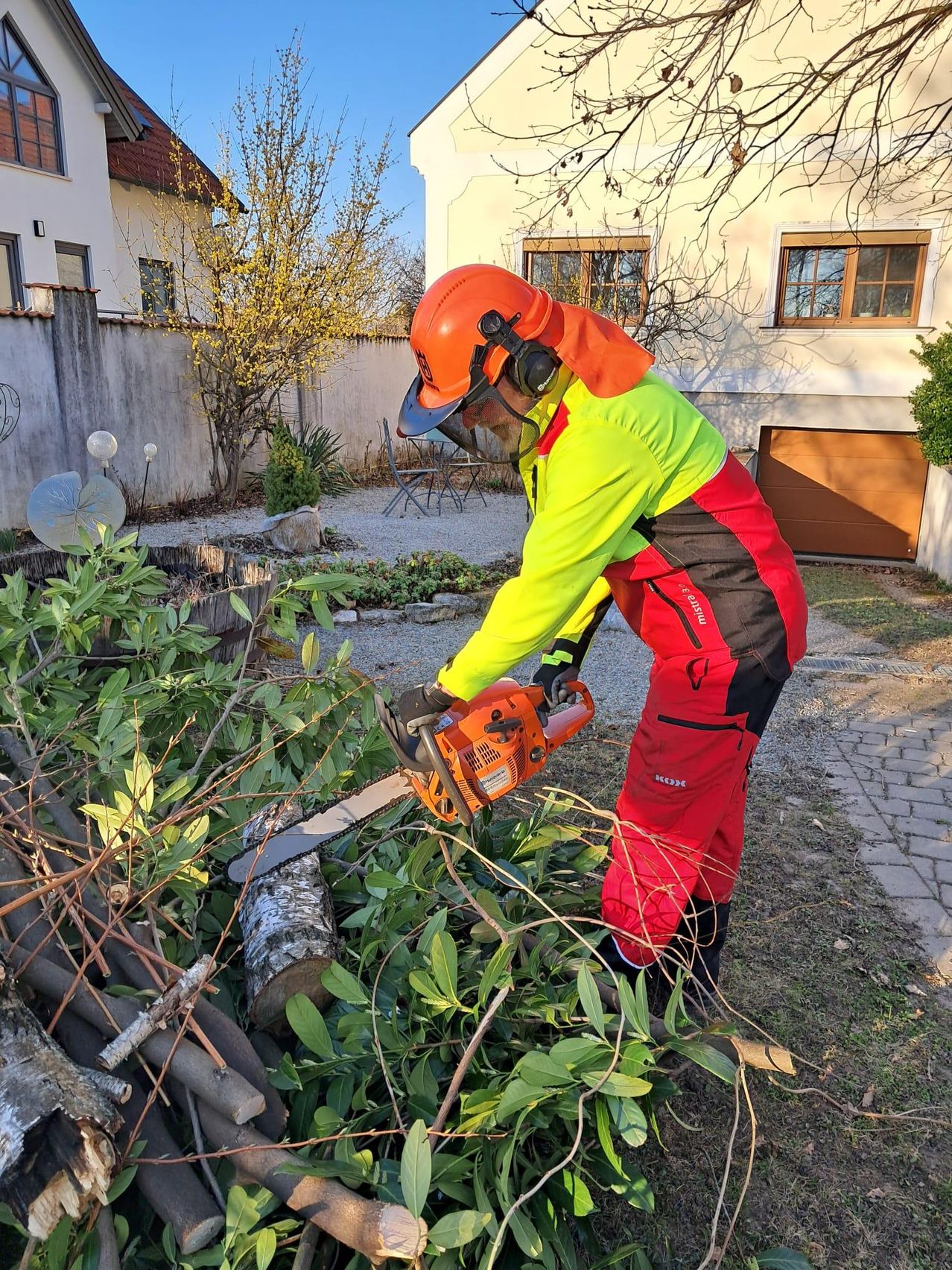 Ein Mann in Schutzausrüstung schneidet mit einer Kettensäge Äste in einem Garten. Er steht auf einem Haufen abgeschnittener Äste, mit einem Haus und einer Garage im Hintergrund.